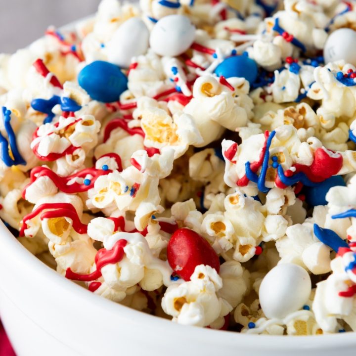 Bowl of red white and blue patriotic popcorn drizzled with red and blue candy melts and topped with M&Ms and sprinkles.