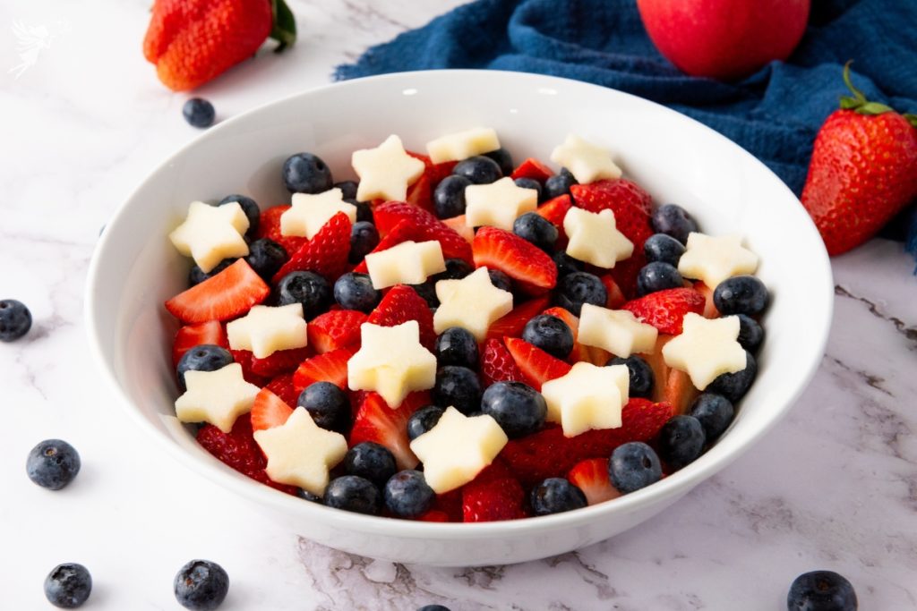 Bowl of star spangled fruit salad with fresh strawberries, blueberries, and star-shaped apple pieces in white serving bowl.