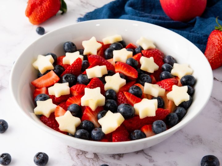 Bowl of star spangled fruit salad with fresh strawberries, blueberries, and star-shaped apple pieces in white serving bowl.