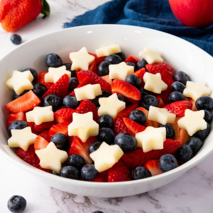 Bowl of star spangled fruit salad with fresh strawberries, blueberries, and star-shaped apple pieces in white serving bowl.