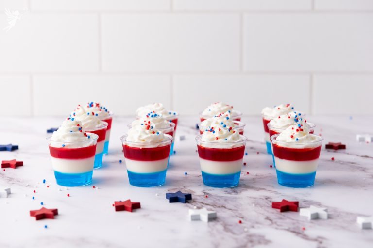 Row of Non Alcoholic Red White & Blue Jello Shots topped with whipped cream and star sprinkles on marble surface with patriotic decorations.