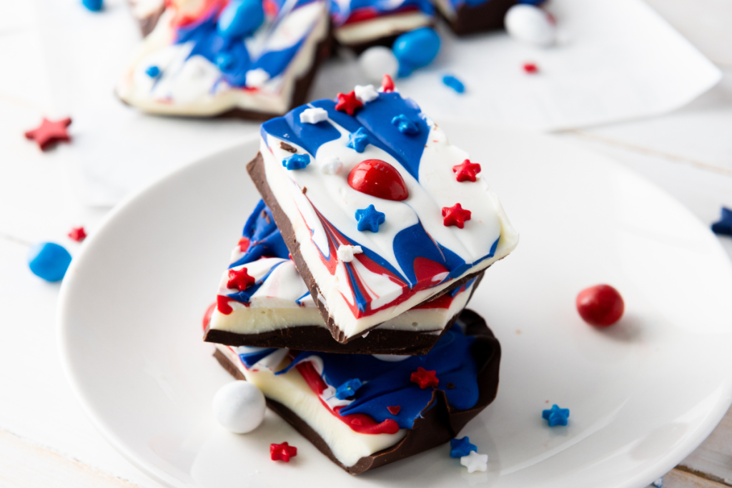 Stack of finished Red White & Blue Chocolate Bark pieces decorated with patriotic swirled candy and star sprinkles on white plate.