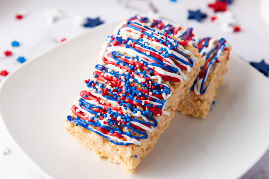 Two finished Patriotic Red White & Blue Rice Krispies bars decorated with candy drizzle and festive sprinkles on white plate.