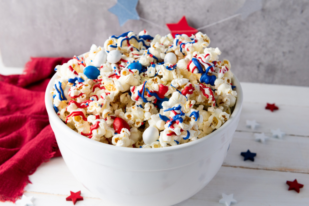 Finished red white and blue patriotic popcorn in white bowl decorated with candy drizzle, M&Ms, and sprinkles with star decorations.