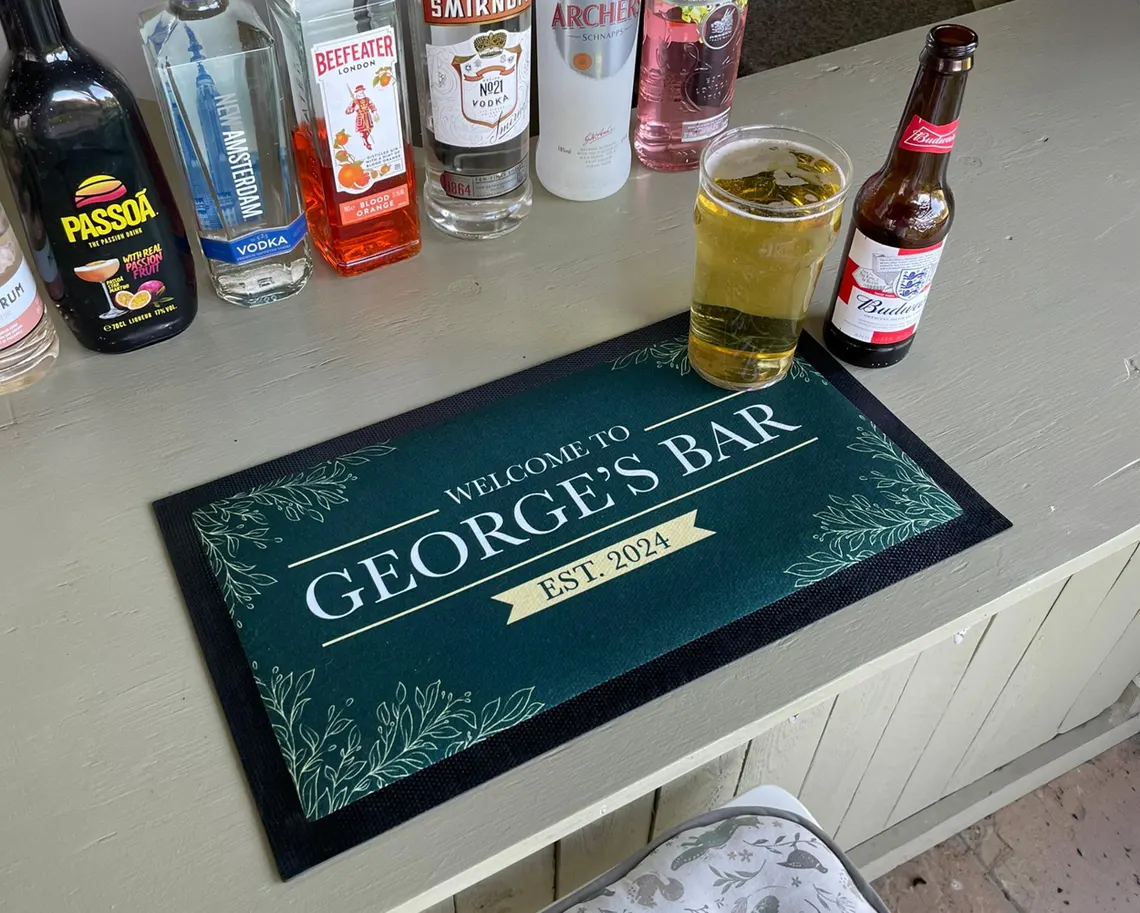 Custom bar mat with "George's Bar" personalization and decorative leaf motifs on home bar setup with various bottles—thoughtful gifts for cocktail lovers. Etsy