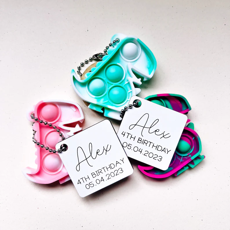 Colorful dinosaur party favors featuring pop-it fidget toys in pink, turquoise, and teal with personalized birthday tags attached via ball chains. Etsy
