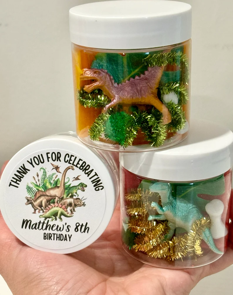 Dinosaur playdough jar dino party favors displayed in transparent containers with dinosaur figurines, glitter decorations, and custom celebration thank-you lid labels. Etsy
