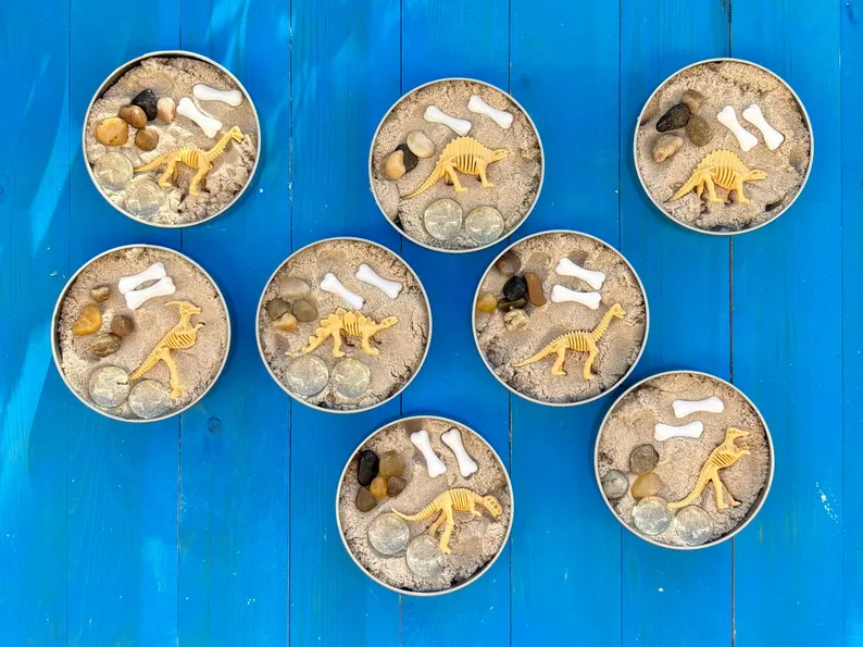 Dinosaurs fossil tin dino party favors featuring clear jars with white lids, filled with colorful dinosaur toys, metallic tinsel, and personalized birthday labels. Etsy