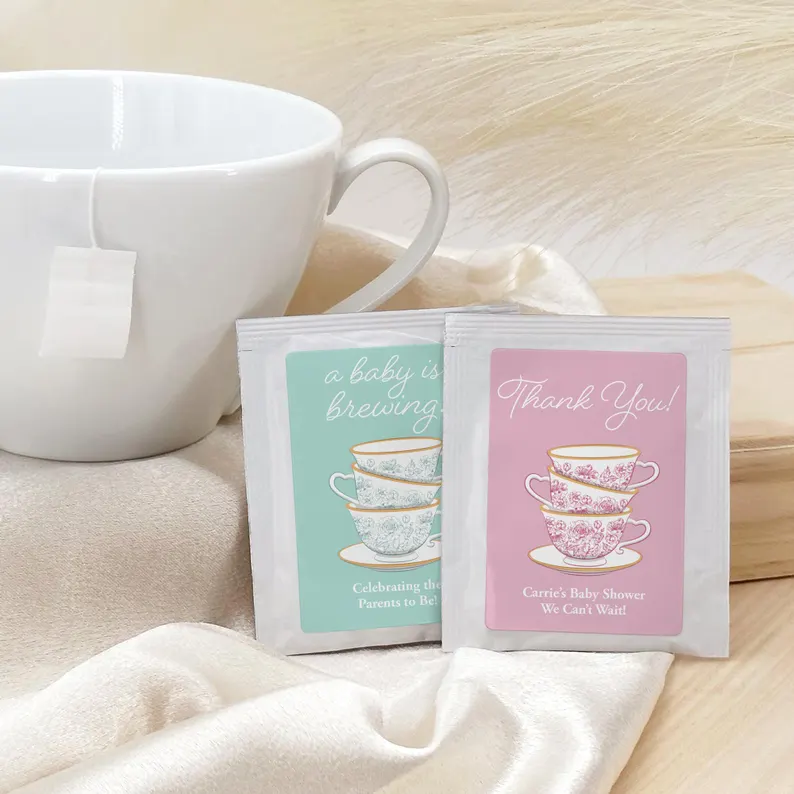 Personalized tea baby shower party favors displaying custom tea packets in mint and pink designs with stacked teacup illustrations beside white mug. Etsy