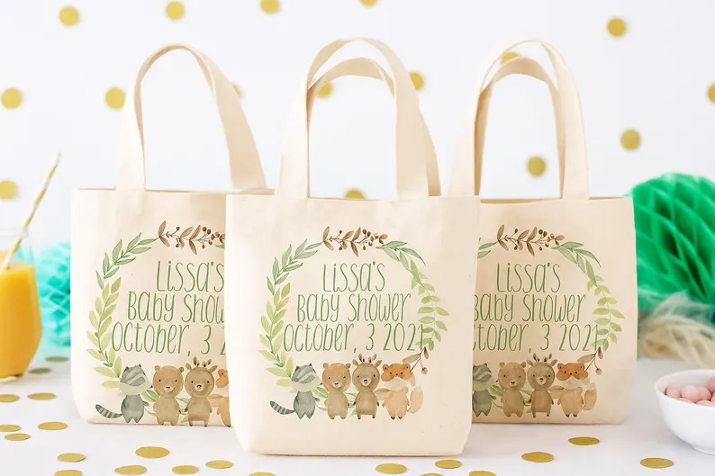 Tote bags baby shower party favors featuring cream canvas bags with woodland animal wreath design and personalized event details on gold polka dot background. Etsy