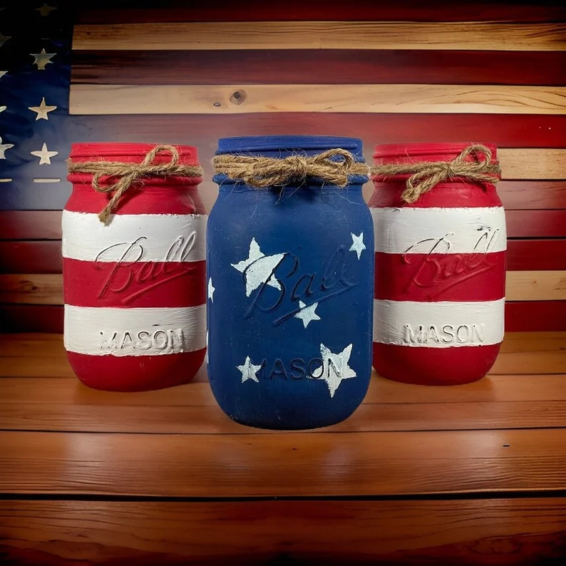 Three hand-painted American flag mason jars in red, white, and blue with twine bows — rustic and charming 4th of July hostess gifts displayed against a wooden flag backdrop. Etsy