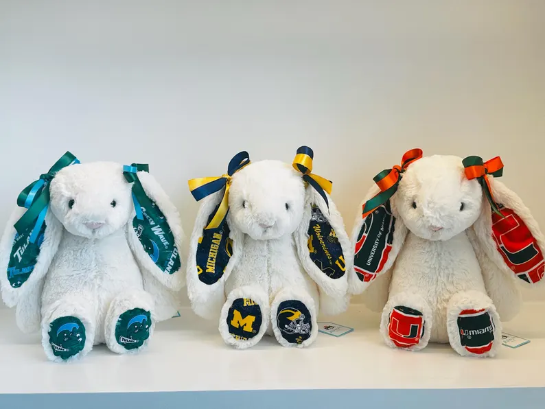 Three white Jellycat bunny plushies customized with university themed ears, bows, and foot patches representing different colleges, among the cutest graduation gifts. Etsy