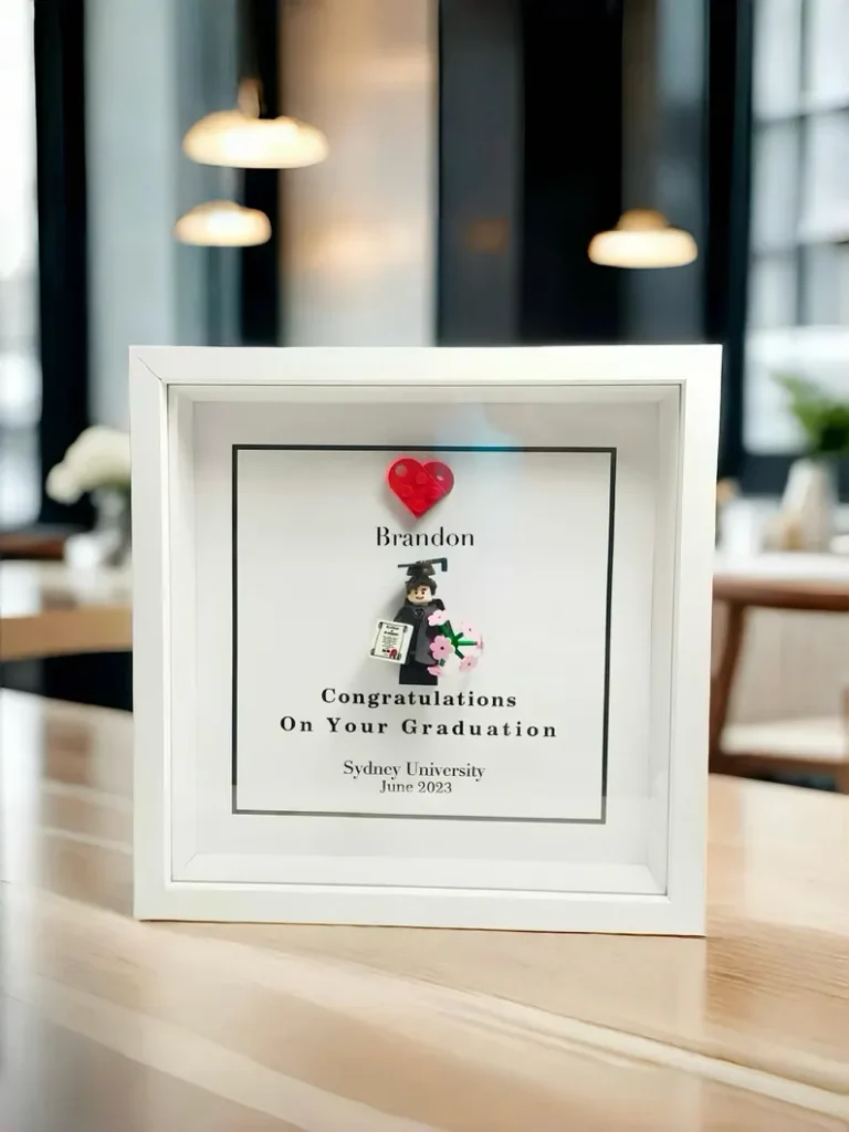 White shadow box frame featuring a personalized LEGO minifigure in graduation gown with a name and university details, one of the most creative graduation gifts. Etsy