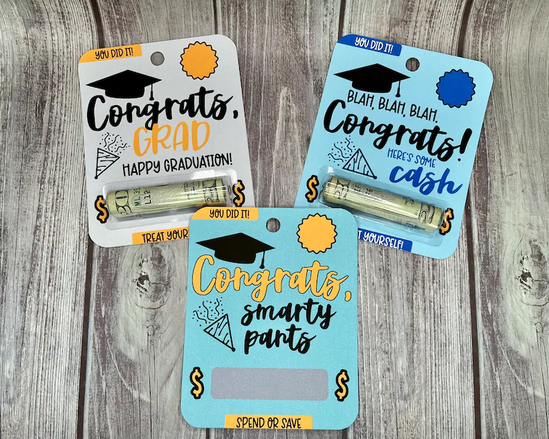 Three colorful printed cards holding rolled cash with funny congratulatory messages, offering a lighthearted and budget friendly option among graduation gifts. Etsy