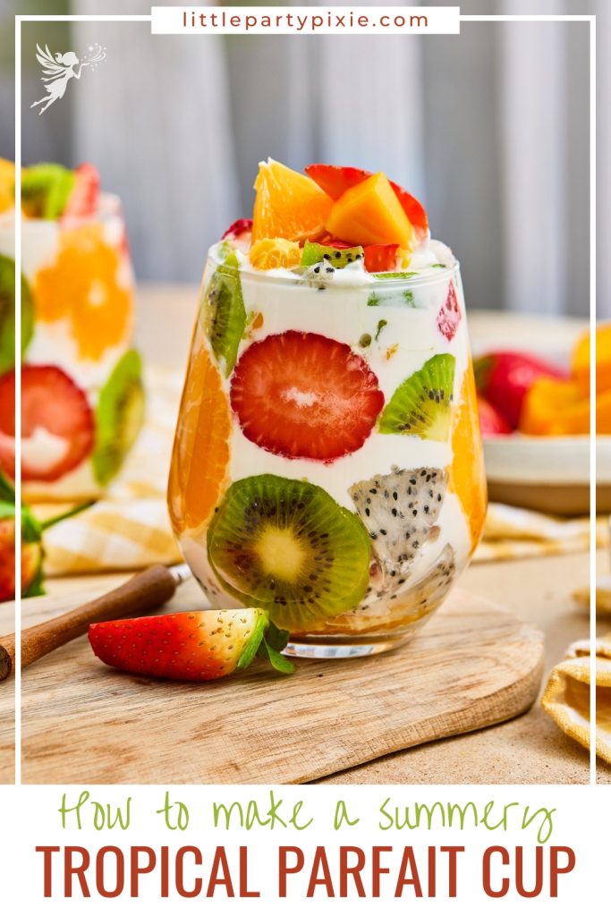 A summery tropical fruit parfait cup filled with colorful layers of kiwi, strawberry, orange, dragon fruit, and whipped cream on a rustic wooden board.