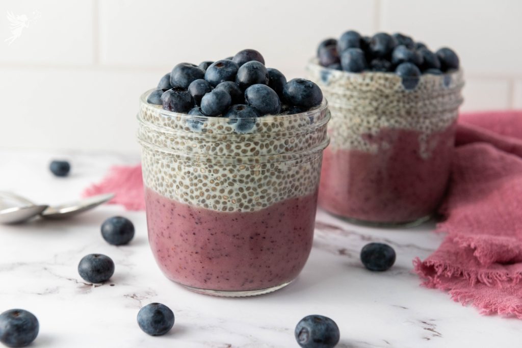 Two blueberry chia pudding cups layered with purple blueberry base, vanilla chia pudding, and fresh blueberries on a marble surface.