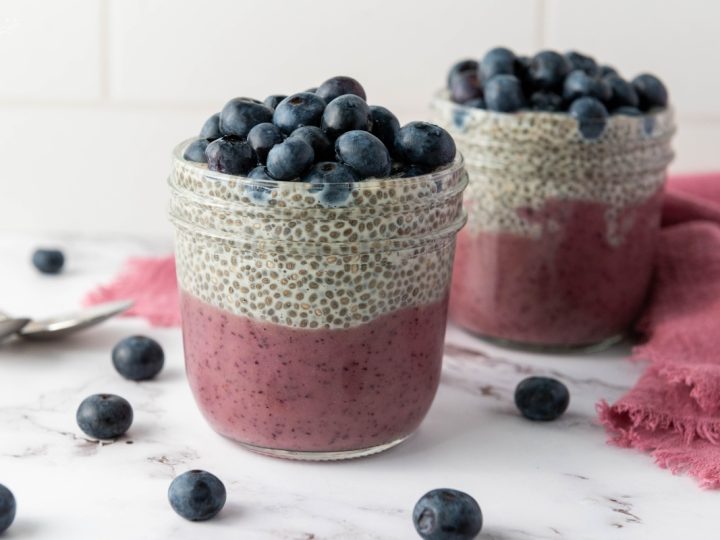 Two blueberry chia pudding cups layered with purple blueberry base, vanilla chia pudding, and fresh blueberries on a marble surface.