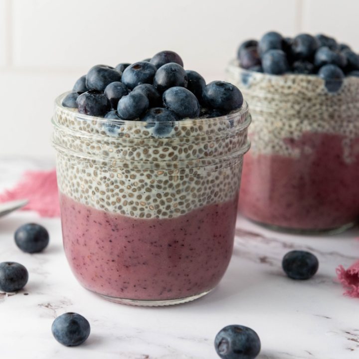 Two blueberry chia pudding cups layered with purple blueberry base, vanilla chia pudding, and fresh blueberries on a marble surface.