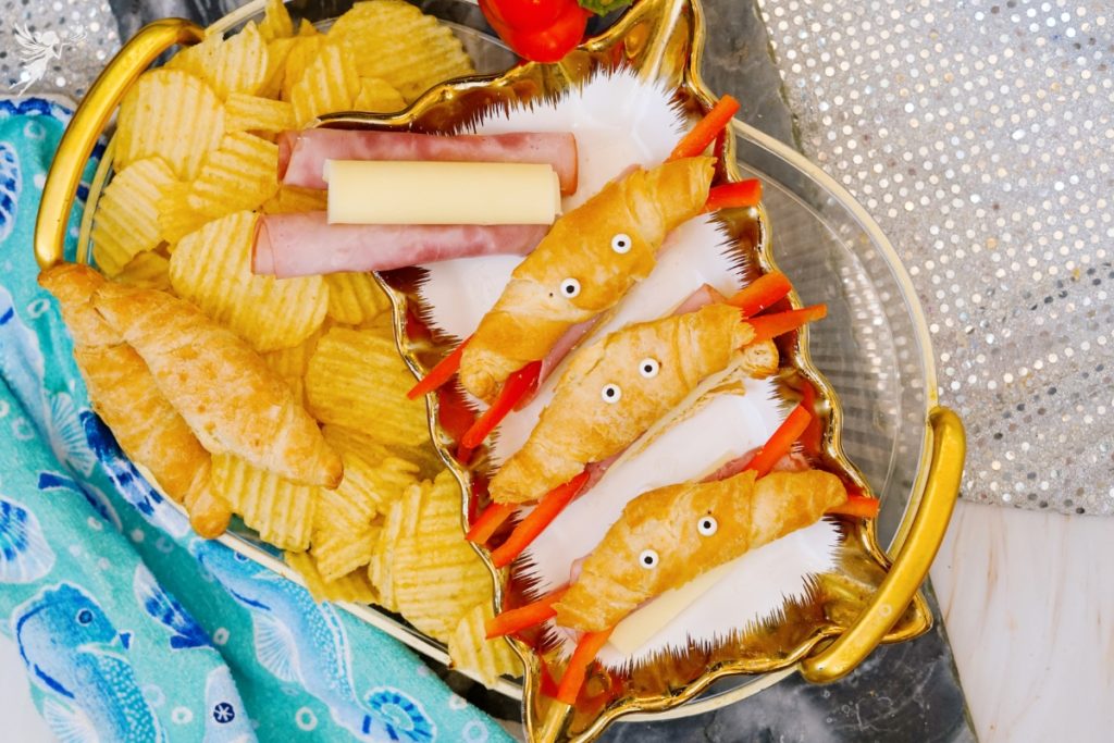 Overhead view of a gold shell shaped serving tray filled with cute crab croissants recipe bites made with mini croissants, string cheese, ham, and red pepper claws alongside ridged potato chips.