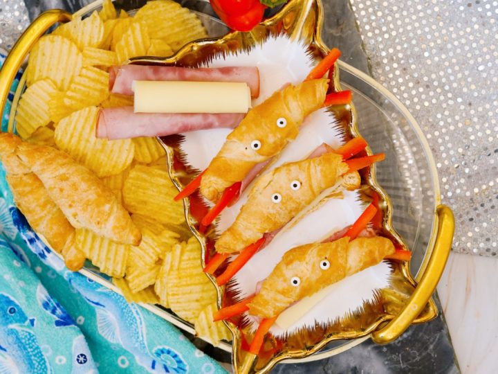 Overhead view of a gold shell shaped serving tray filled with cute crab croissants recipe bites made with mini croissants, string cheese, ham, and red pepper claws alongside ridged potato chips.