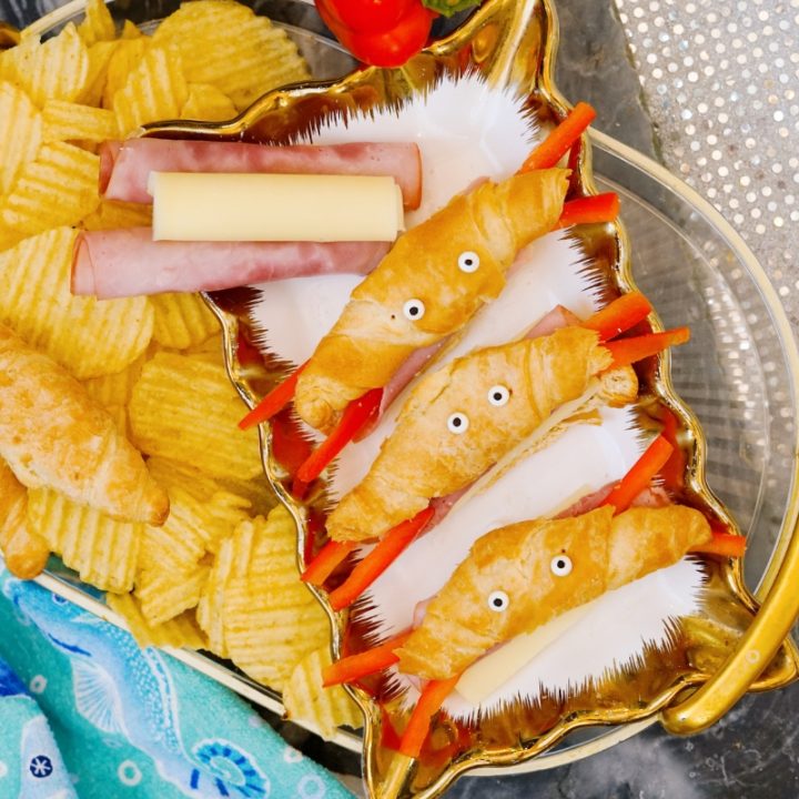 Overhead view of a gold shell shaped serving tray filled with cute crab croissants recipe bites made with mini croissants, string cheese, ham, and red pepper claws alongside ridged potato chips.