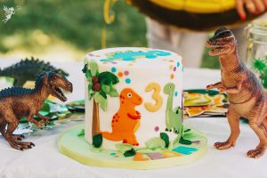 dino party themed cake with cute dinosaurs cut out in fondant and number 3 surrounded by plastic dino decorations