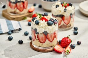 Red white and blue berry trifles served in clear glasses with layered strawberries, blueberries, and whipped cream on a wooden coaster.
