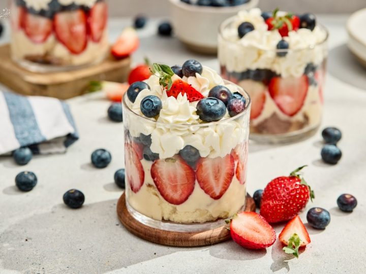 Red white and blue berry trifles served in clear glasses with layered strawberries, blueberries, and whipped cream on a wooden coaster.