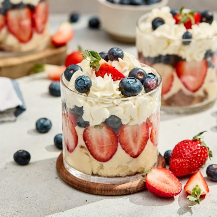 Red white and blue berry trifles served in clear glasses with layered strawberries, blueberries, and whipped cream on a wooden coaster.