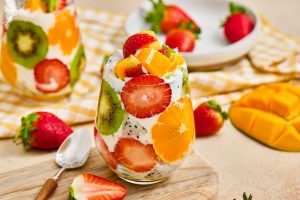 Vibrant tropical fruit parfait cups layered with fresh strawberries, kiwi, orange slices, mango, and creamy yogurt served on a wooden board.