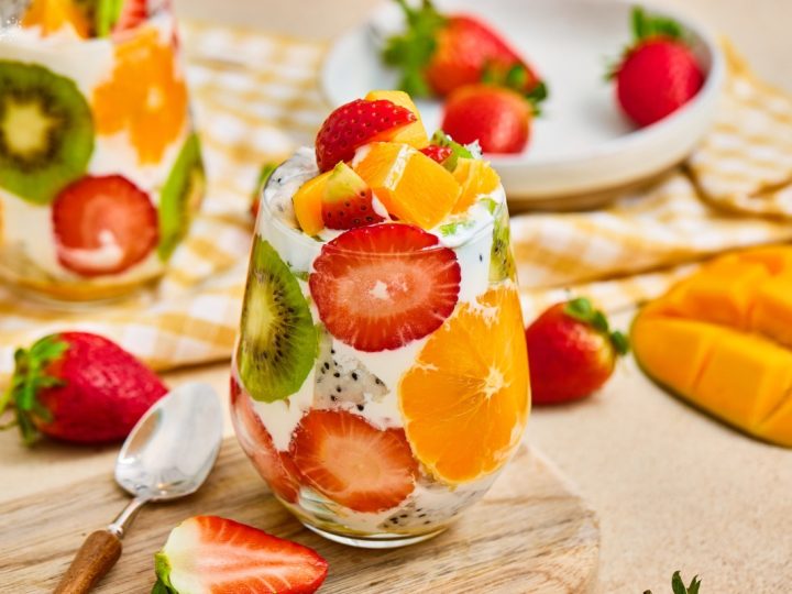Vibrant tropical fruit parfait cups layered with fresh strawberries, kiwi, orange slices, mango, and creamy yogurt served on a wooden board.