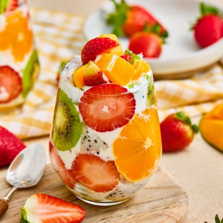 Vibrant tropical fruit parfait cups layered with fresh strawberries, kiwi, orange slices, mango, and creamy yogurt served on a wooden board.