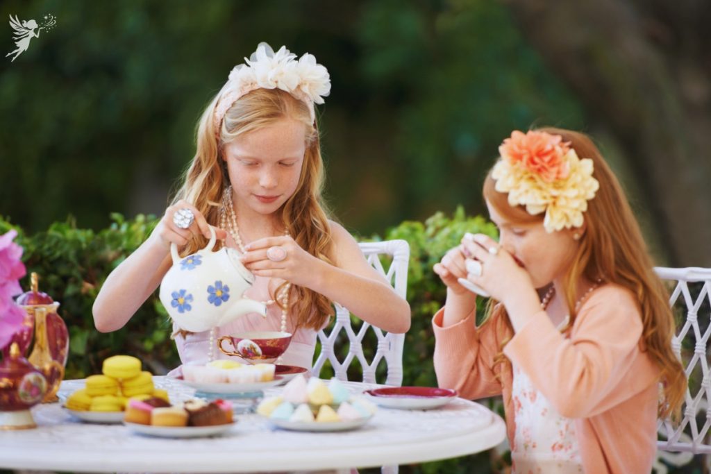 two young girls dressed up in fancy clothing having a tea party in the garden pouring and drinking tea in cups