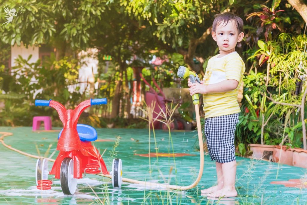 a small child holding a hose in the garden washing his tricycle