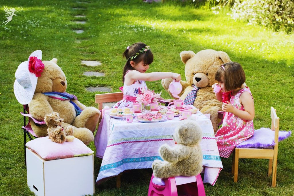 two small sitting with giant teddy bears serving them tea and drinking from palstic cups