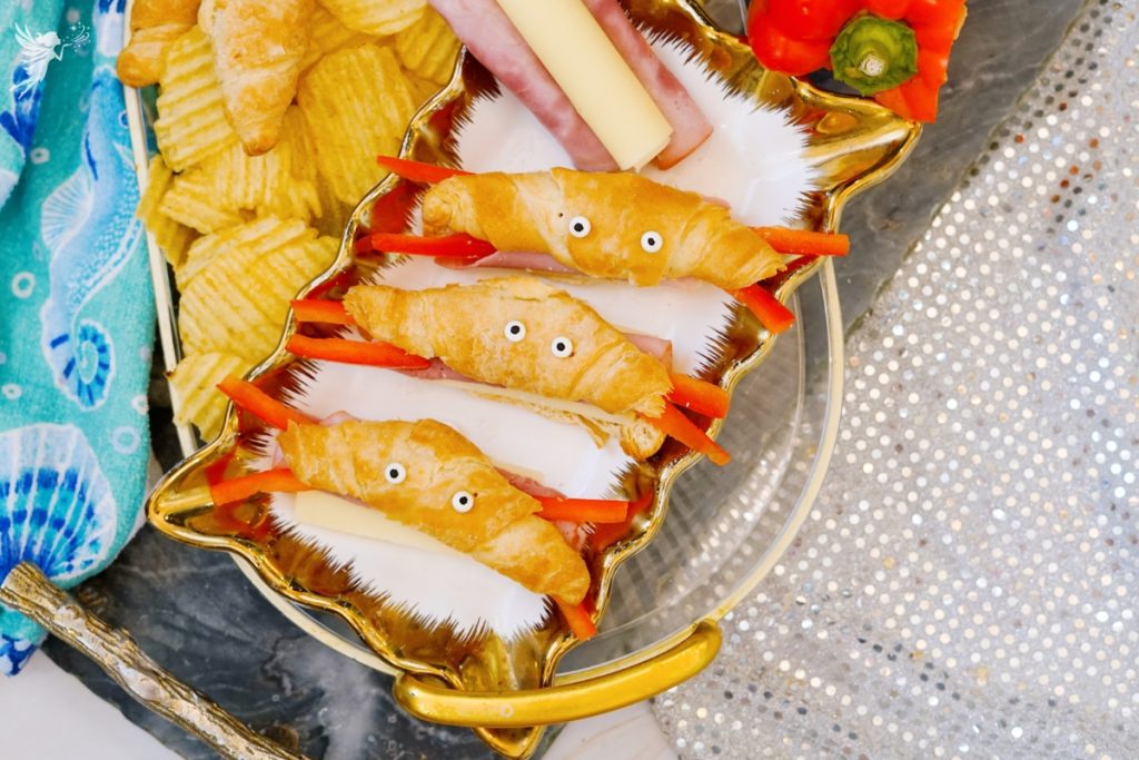 Close up of finished crab croissants recipe arranged on a decorative gold shell tray with string cheese bodies, red pepper claws, candy eyes, and ridged chips as a fun party snack.