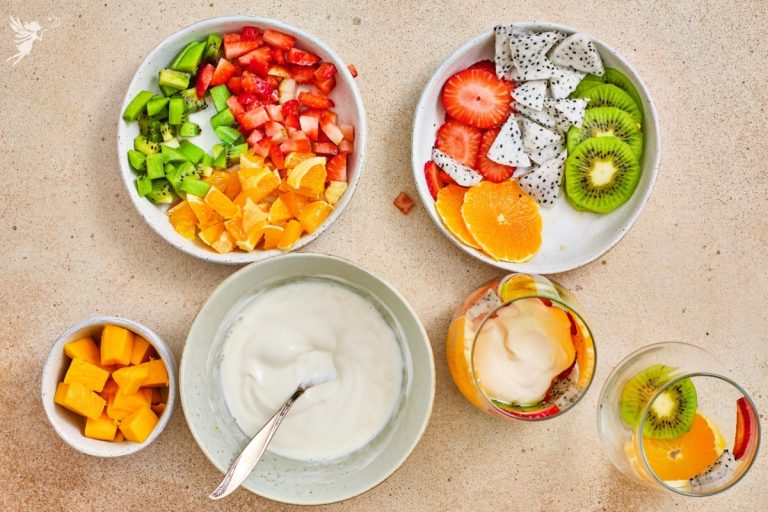 Flat lay of tropical fruit parfait cups ingredients including chopped kiwi, strawberries, dragon fruit, orange segments, mango cubes, and creamy yogurt in bowls.