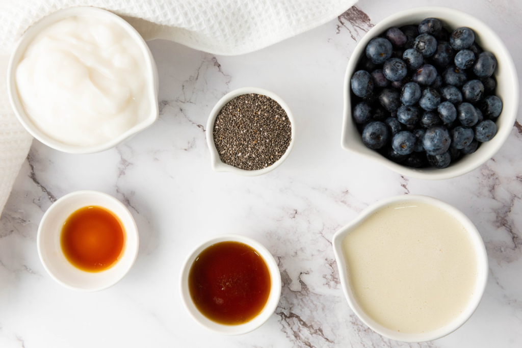 Ingredients for Blueberry Chia Pudding Cups including chia seeds, plant-based milk, yogurt, vanilla, maple syrup, and fresh blueberries arranged on a marble surface.