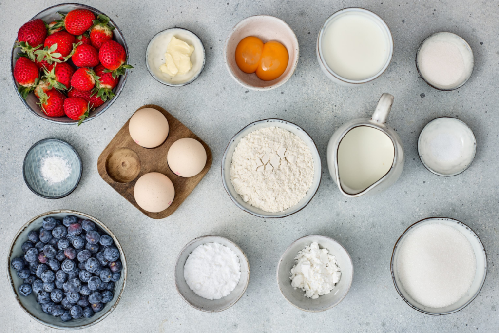 Overhead flat lay of all ingredients for red white and blue berry trifles including fresh strawberries, blueberries, eggs, flour, milk, butter, and sugar.