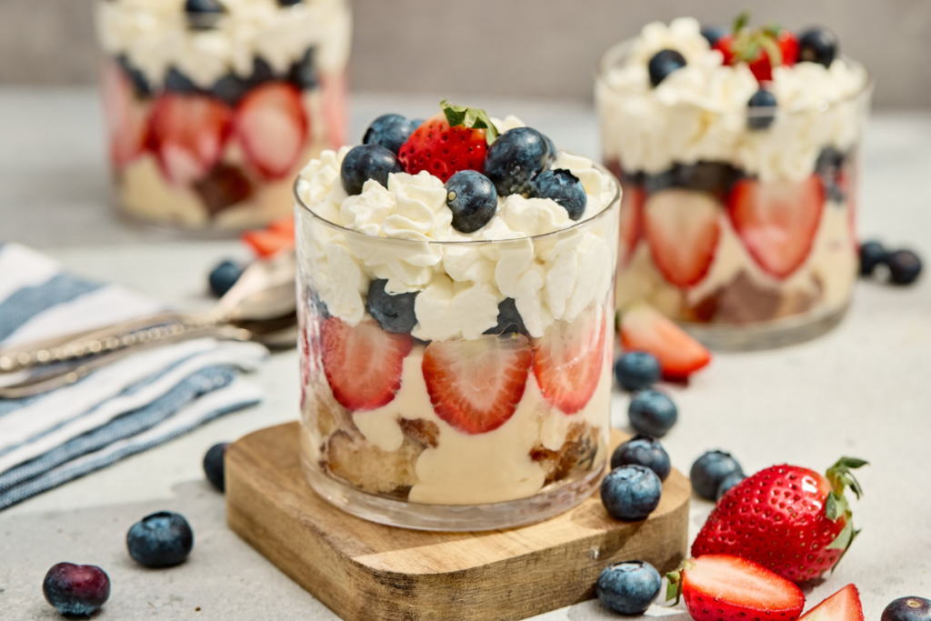 Three finished red white and blue berry trifles in clear glasses layered with sponge cake, vanilla pudding, fresh strawberries, blueberries, and whipped cream.