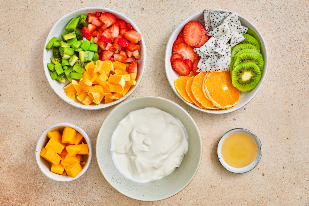 Overhead view of all ingredients needed to make tropical fruit parfait cups including sliced kiwi, strawberries, dragon fruit, orange, mango cubes, yogurt, and honey.