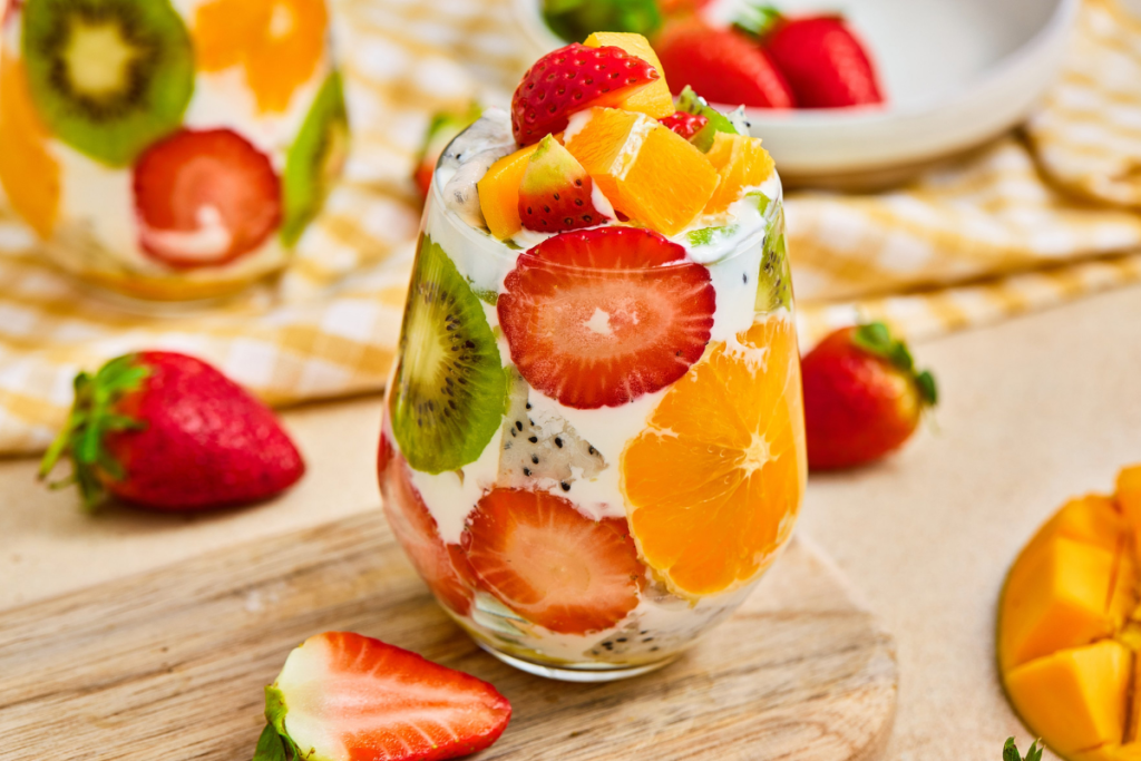 A finished tropical fruit parfait cup filled with vibrant layers of strawberry, kiwi, orange, dragon fruit, mango, and yogurt on a rustic wooden board.