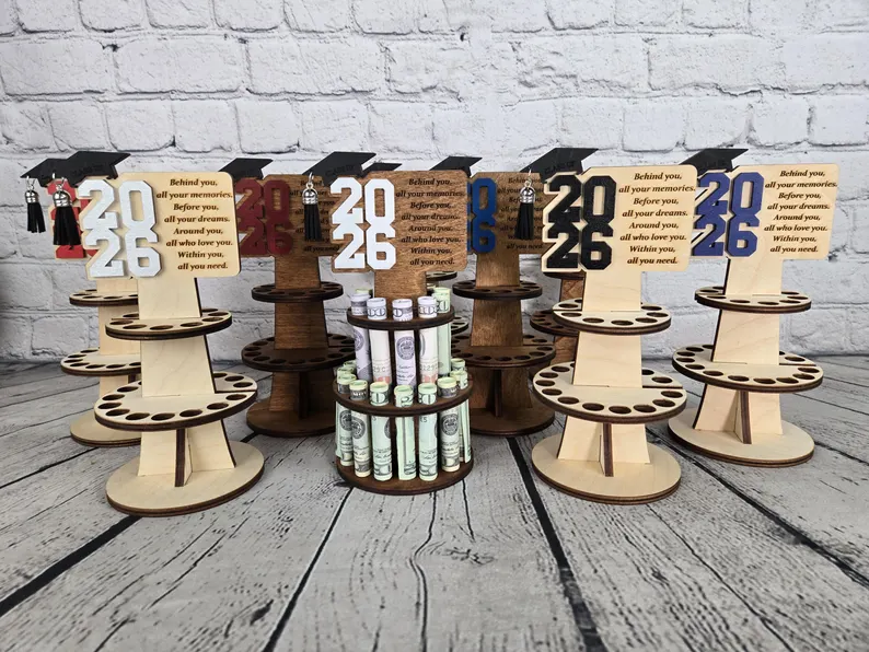 Multiple tiered wooden stands displaying rolled cash bills with "2026" graduation cap cutouts in various colors, among the most festive graduation gifts. Etsy