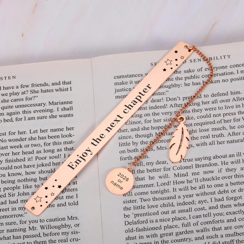 Rose gold metal bookmark engraved with "Enjoy the next chapter" and a feather charm, resting open on a book, among the most elegant graduation gifts. Etsy