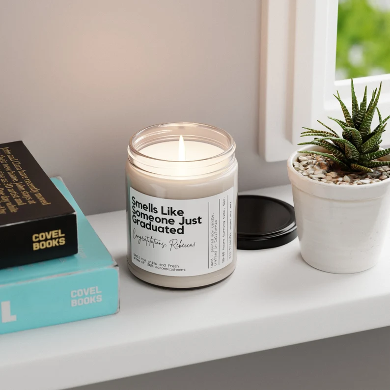 Lit soy candle in a glass jar labeled "Smells Like Someone Just Graduated," displayed on a windowsill beside books, among the wittiest graduation gifts. Etsy
