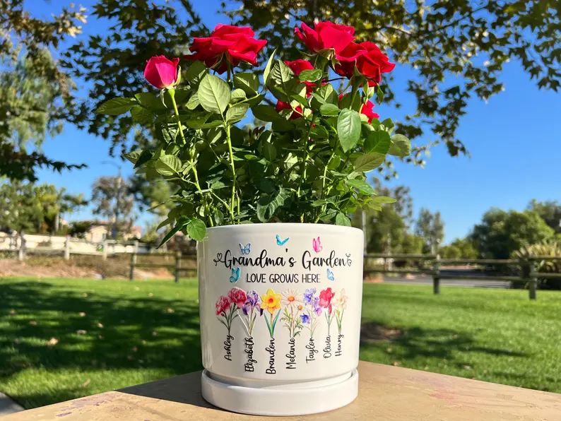 White ceramic flower pot with Grandma's Garden text featuring colorful birth flowers and grandchildren names holding red roses, beautiful grandma gifts from grandkids.