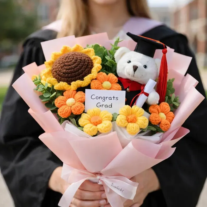 A graduate in a black gown holds a pink-wrapped bouquet of crocheted sunflowers and daisies with a tiny teddy bear wearing a graduation cap, making it one of the most charming graduation gifts. Etsy