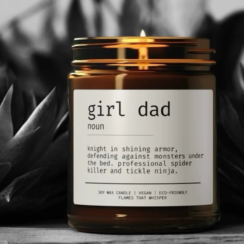 A lit amber jar soy wax candle labeled "girl dad" with a humorous definition, serving as a fun and lighthearted first Father's Day gift idea for girl dads. Etsy
