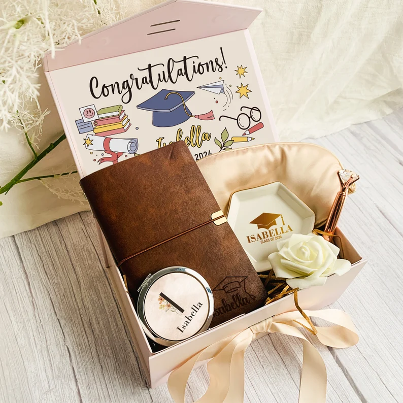 An open pink gift box filled with a leather journal, compact mirror, personalized ring dish, and a rose, beautifully curated as thoughtful and elegant graduation gifts for her. Etsy