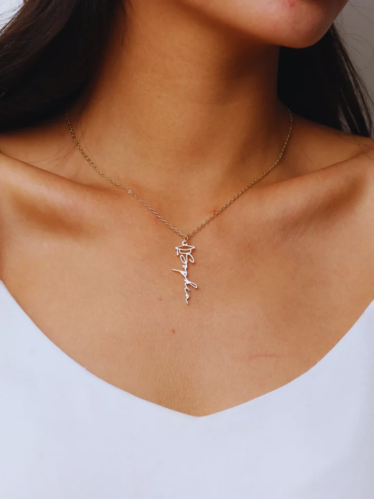 A woman wearing a delicate gold chain necklace featuring a custom script name pendant topped with a graduation cap charm, a meaningful and wearable piece among graduation gifts for her. Etsy